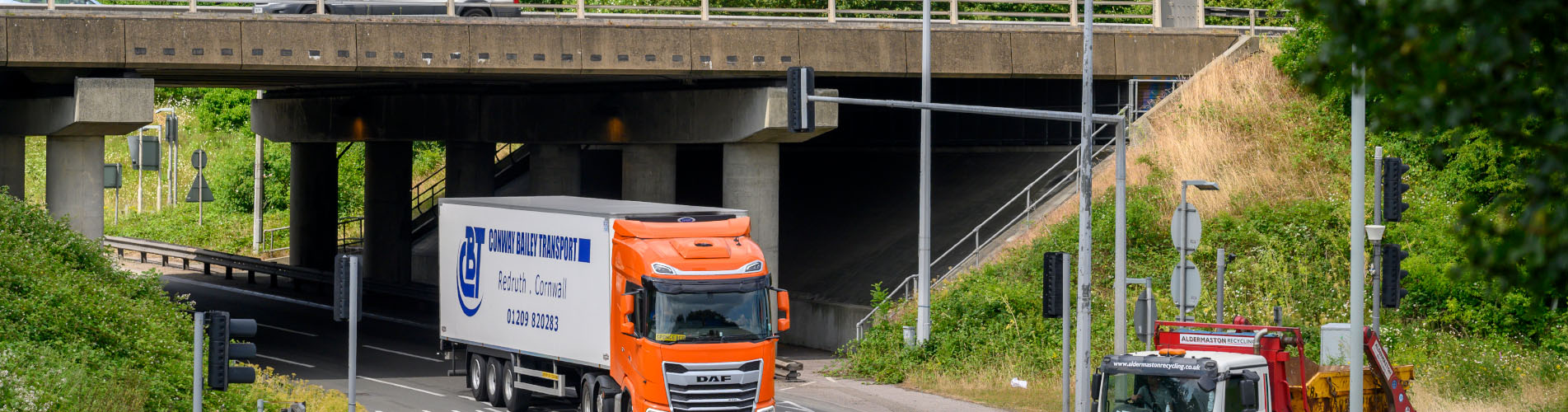 A three-lane traffic light roundabout with some traffic including HGVs with an overpass for through traffic. There is a background of trees and greenery against a cloudy sky.