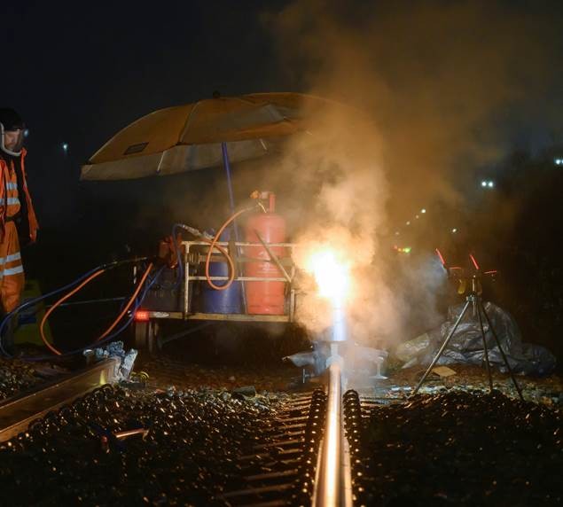 Night-time railway maintenance activity showing equipment and a bright welding flame on the track, with smoke rising and a worker in high-visibility orange protective clothing standing nearby.