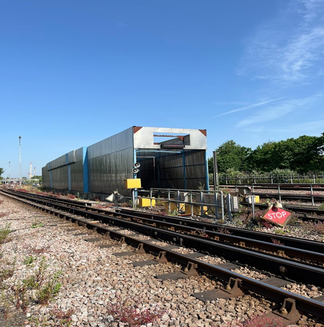 A metal rain wash structure at a railway depot against a blue sky background, with railway tracks running in front and behind the train wash.