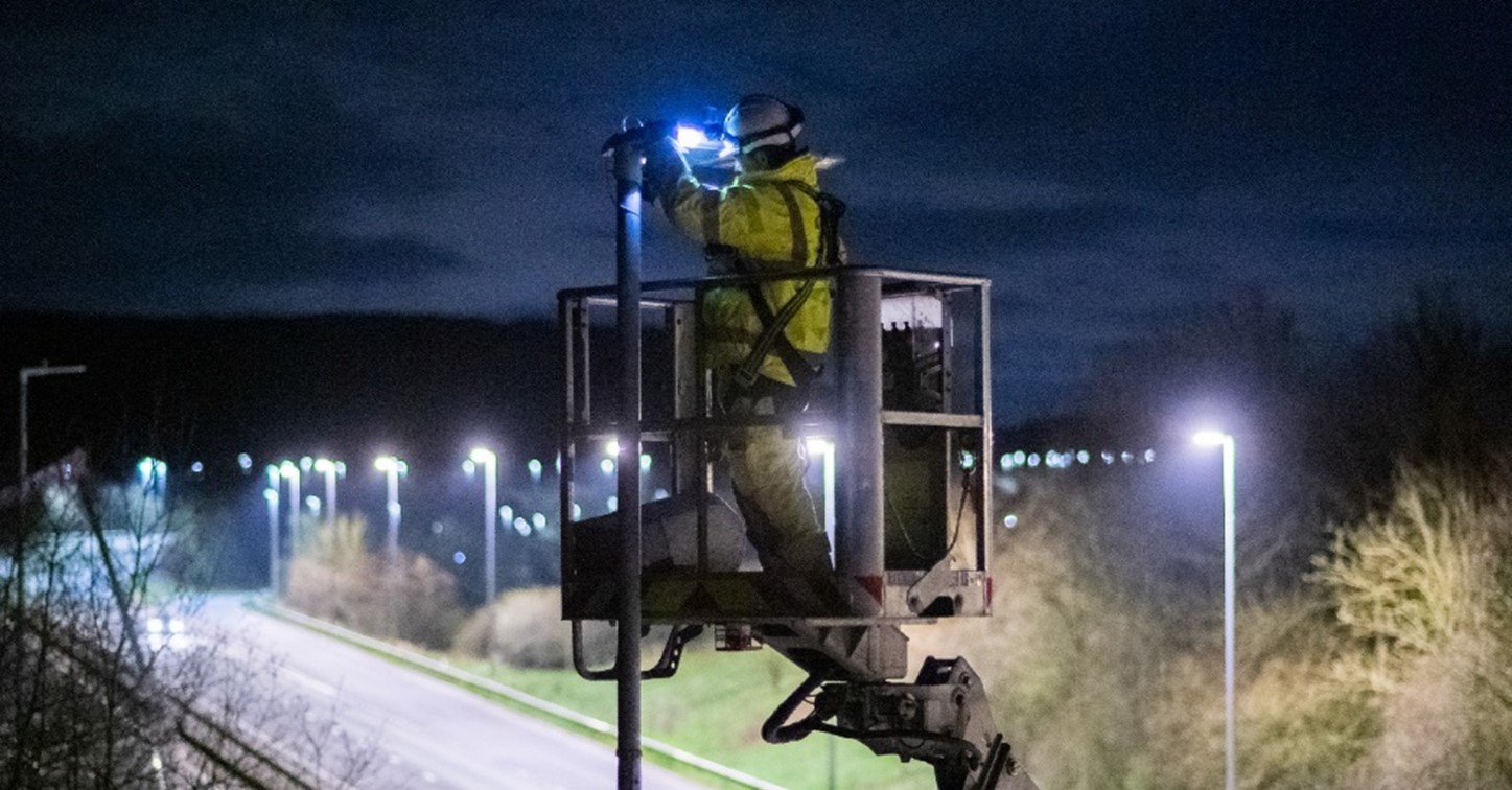 A picture taken at night of a highway worker on a cherry picker replacing a bulb in a streetlight alongside the SRN