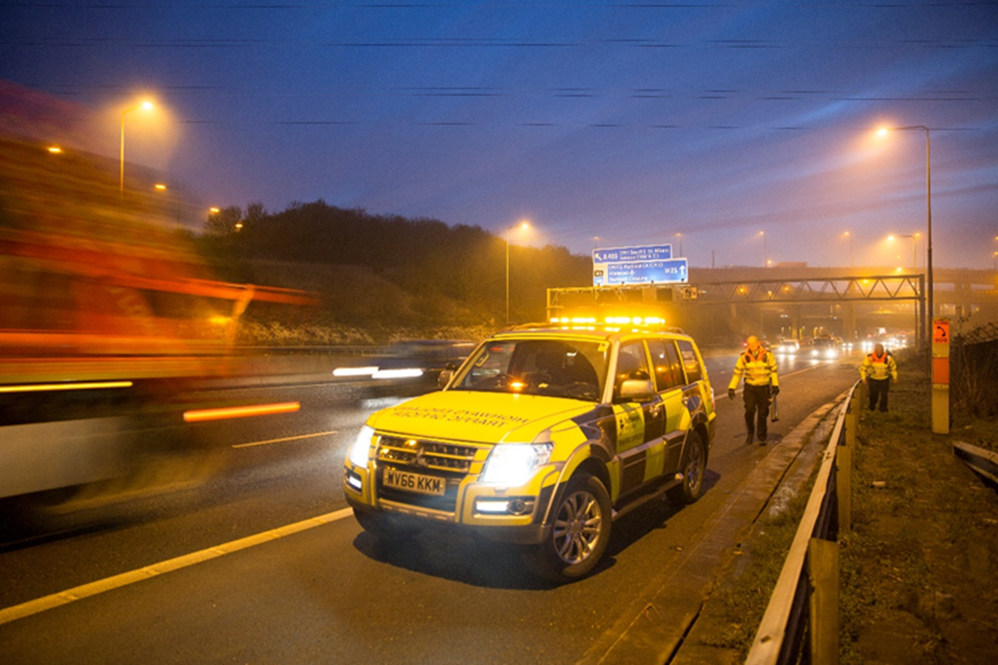 A picture taken at night of a National Highway traffic officer vehicle parked on the hard shoulder with traffic driving past whilst two traffic officers are returning to their vehicle