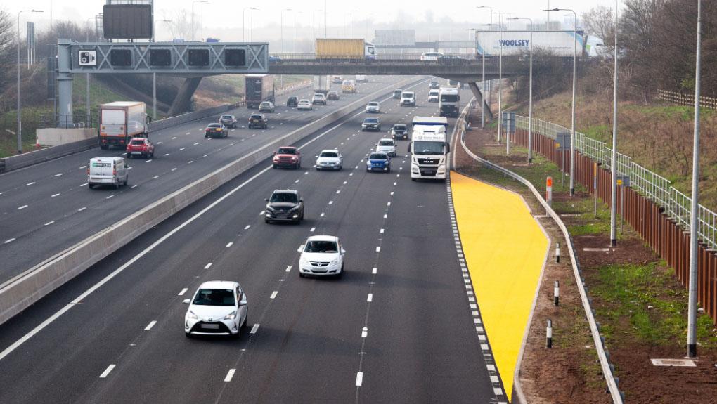 A view over a section of the road near a yellow lay-by with a bridge in the background.