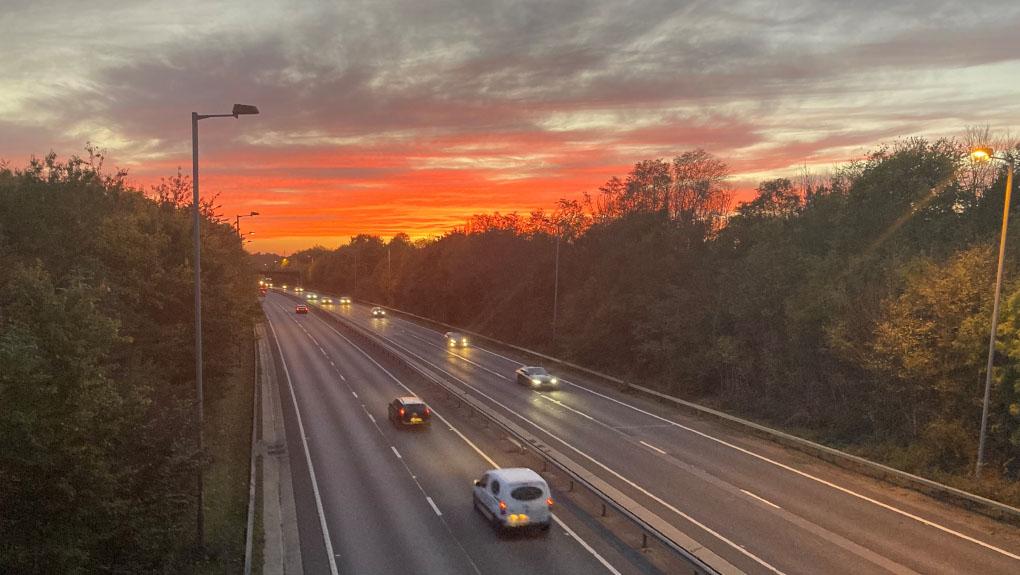 A picture of a dual carriageway bordered by tall trees with a sunset on the horizon