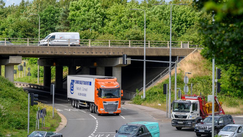 A three-lane traffic light roundabout with some traffic including HGVs with an overpass for through traffic. There is a background of trees and greenery against a cloudy sky.
