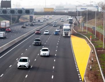 A view over a section of the road near a yellow lay-by with a bridge in the background.