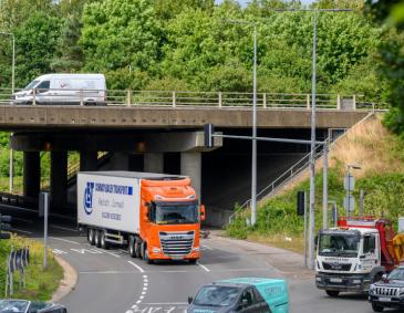 A three-lane traffic light roundabout with some traffic including HGVs with an overpass for through traffic. There is a background of trees and greenery against a cloudy sky.